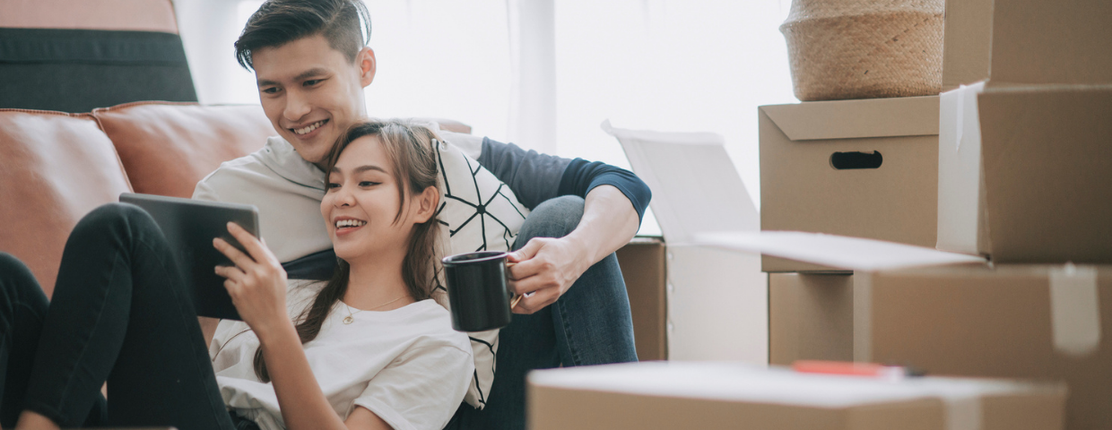 A man and a woman, surrounded by moving boxes, sit on the couch looking at a tablet.