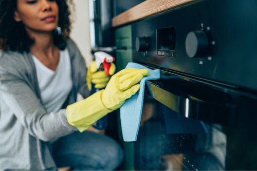A woman wearing rubber gloves cleans and disinfects the handle of her oven range as she cleans the kitchen