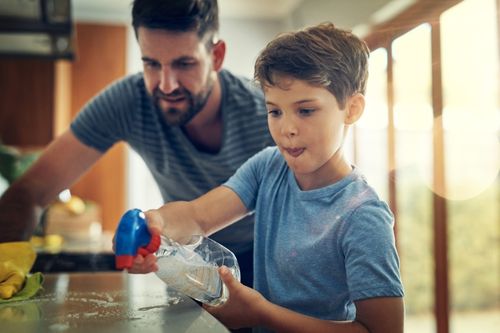 A father and his young son work together to clean the kitchen, spraying the countertops with an all-purpose cleaning solution