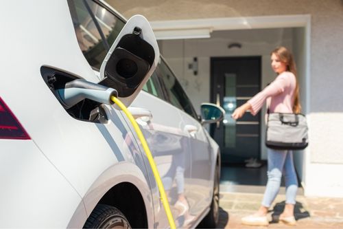 A woman gets home from work and charges her electric vehicle in her driveway just outside the garage.