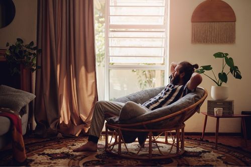 A young man relaxes in the living room of his rental looking out his window with beige curtains and house plants around him
