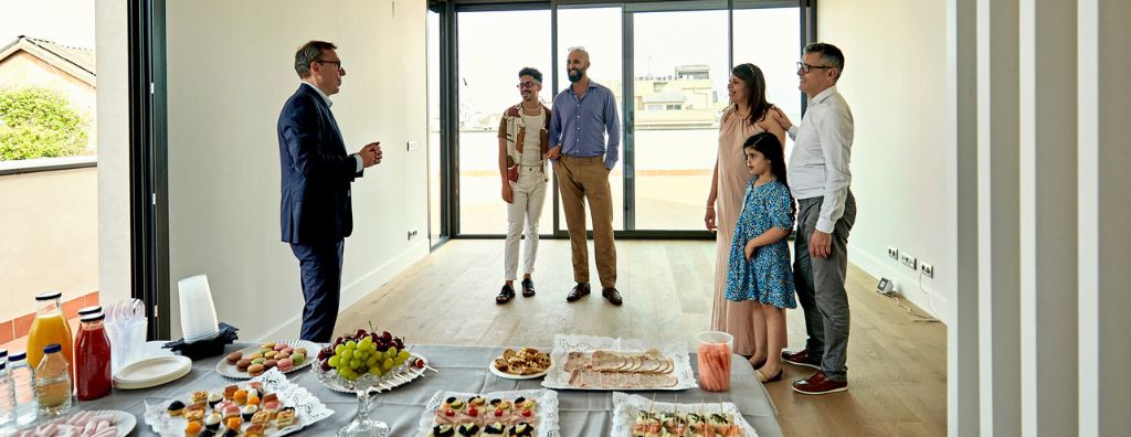 A real estate agent hosts an open house in a new construction home. Two families of prospective buyers listen to him describe the home’s features. There are platters of snacks on a table in the living room.
