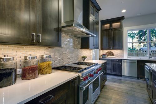 The interior of a modern home kitchen with granite countertops, hardwood floors, dark oak cabinets, and a stainless-steel range and dishwasher.