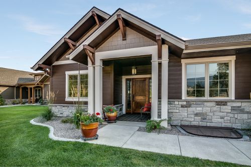 A wide shot from the street of a home with great curb appeal. It is a single-family home in a suburban neighborhood with brown siding, large windows, stone masonry, and thick green grass in the front yard.