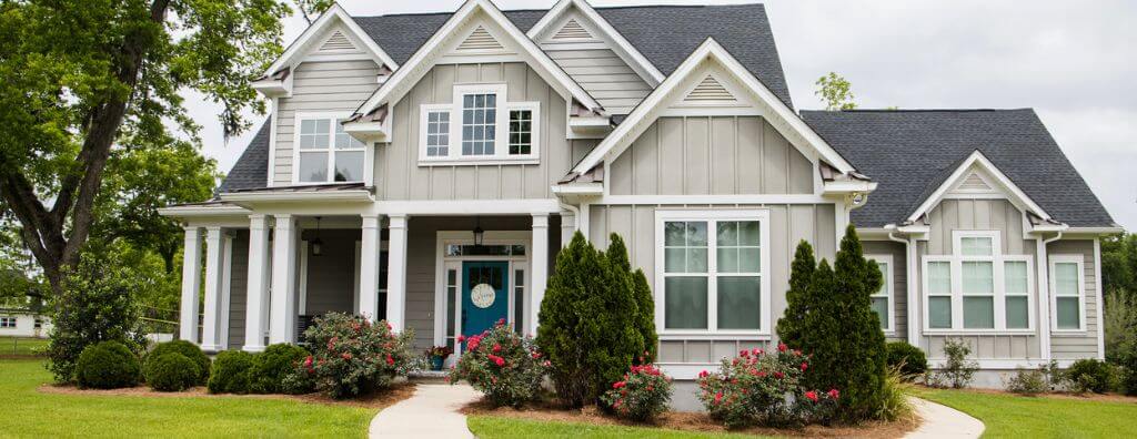 A wide shot from the street of a home with great curb appeal. It is a single-family home in a suburban neighborhood with porch columns, white trim, and grey siding. The flower beds are full of well-landscaped shrubs and rose bushes.