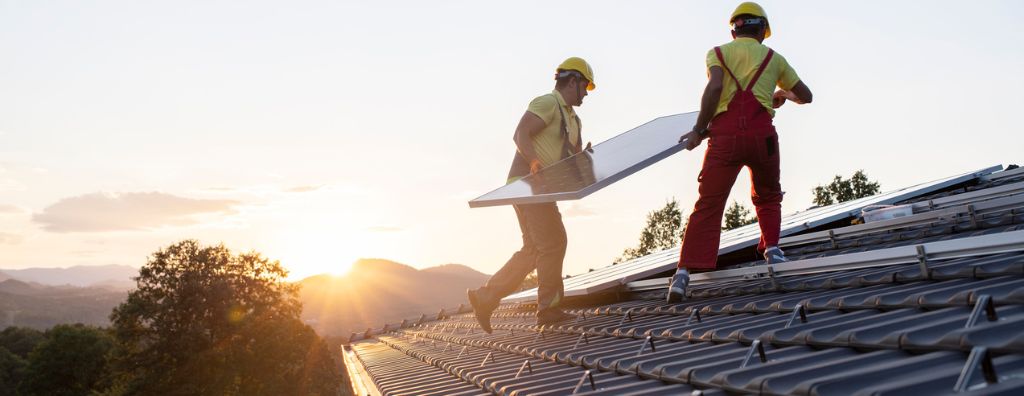 Two male technicians install solar panels on a roof at sunset.