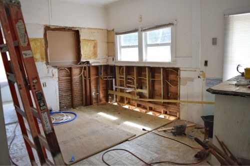 The interior of a fixer-upper house with ripped out kitchen cabinets, drywall, and flooring. The kitchen is being completely gutted and remodeled.