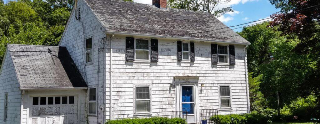 The exterior of a fixer-upper house with deteriorating white siding. The paint is chipped, the roof shows signs of damage, and the window shutters are cracked and uneven.