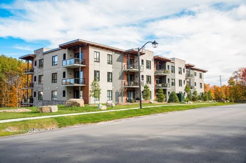 A three-story condominium housing development with gray siding and wood accents. Each condo unit has a small balcony with glass panes extending from the corners of the building. The units share a grassy yard surrounding the building.