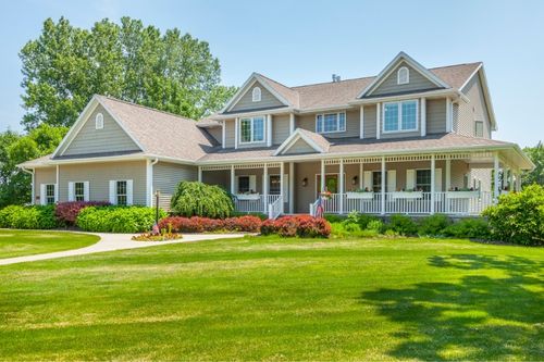 A single-family two-story house with a large front yard, a covered wraparound porch, and an attached garage/bonus space. There are several windows lining the first and second floors and a white fence lines the porch.