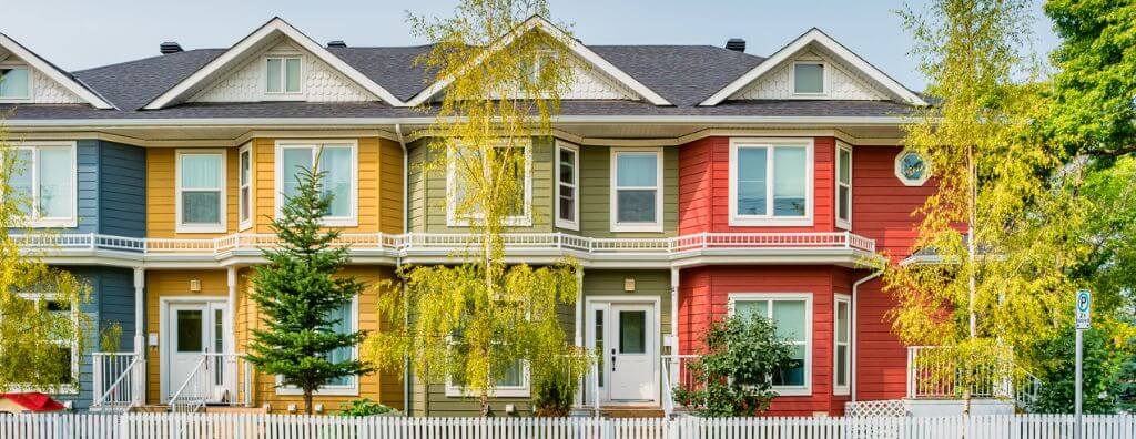 A row of colorful three-story townhouses—blue, yellow, green, and red—with a white picket fence from end to end and a couple trees on each of the four properties.