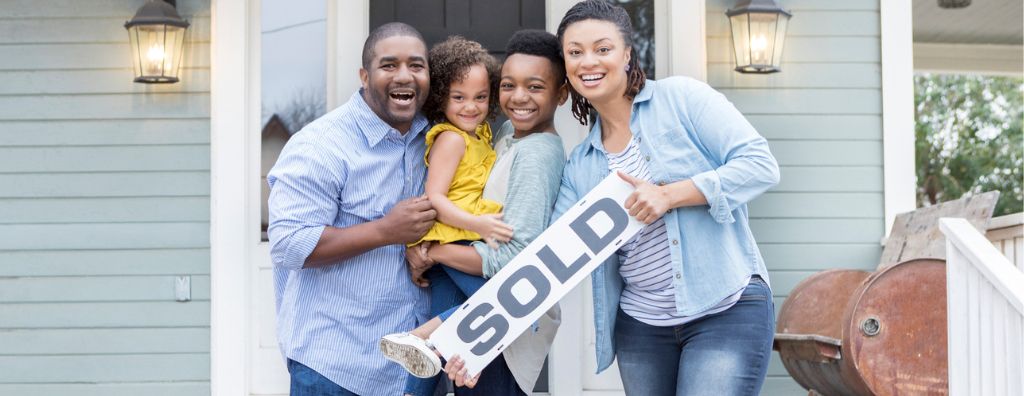 A father, mother, and their two children stand on the front porch of their new home, smiling as they hold a “sold” sign