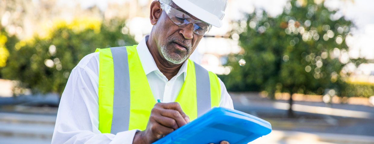 A home inspector wearing a hard hat fills out paperwork on his clipboard.