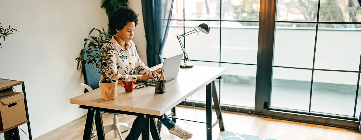 A woman sits in her home office typing on her laptop with houseplants and writing utensils on her desk.