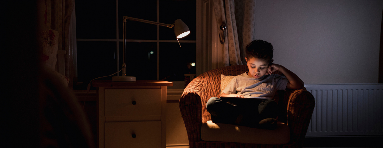 A child sits in a large chair under a lamp reading a book.