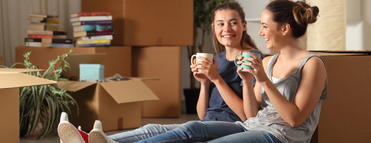 Two young women sit in their new home surrounded by boxes.