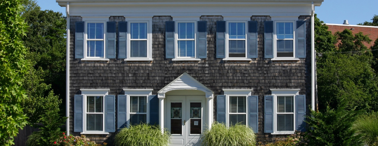 A brown cape cod home surrounded by hedges and trees.