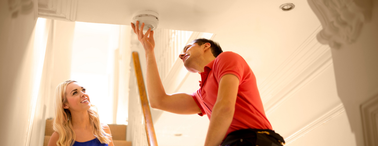 A man and a woman examine a smoke detector in their home.