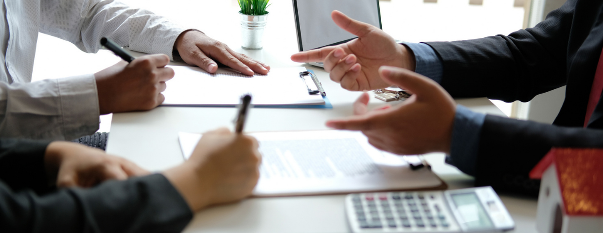 A real estate agent sits at a desk with two people selling their home. All three examine paperwork.