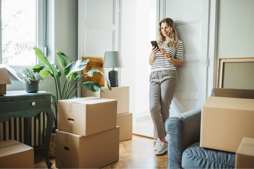 A middle-aged Caucasian woman explores quotes from moving companies on her phone in her living room. She is surrounded by moving boxes.