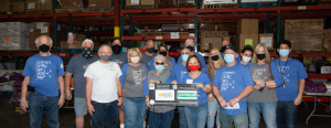 A group of people wearing sanitary masks stand together in a food bank warehouse holding a donation check.
