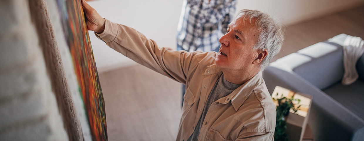 A man hangs a piece of art on a wall while a woman behind him gives him instructions.