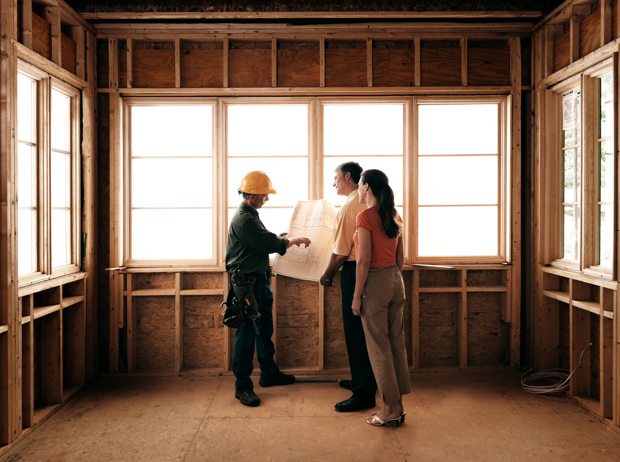 A man and woman look at blueprint plans with a contractor inside a room that’s being remodeled.