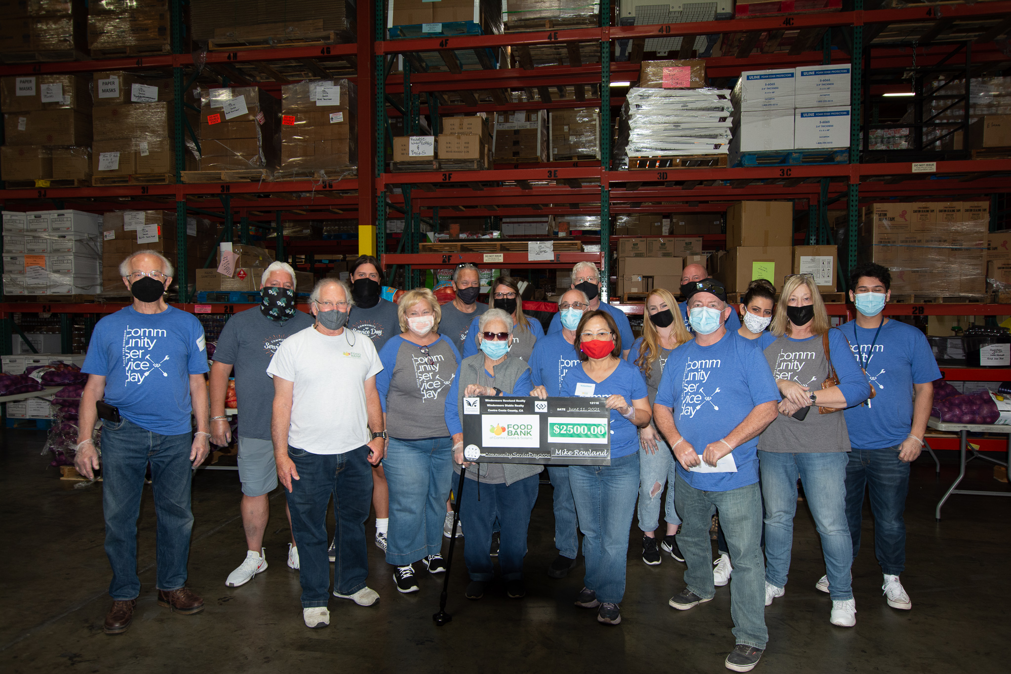 A group of people wearing sanitary masks stand together in a food bank warehouse holding a donation check.