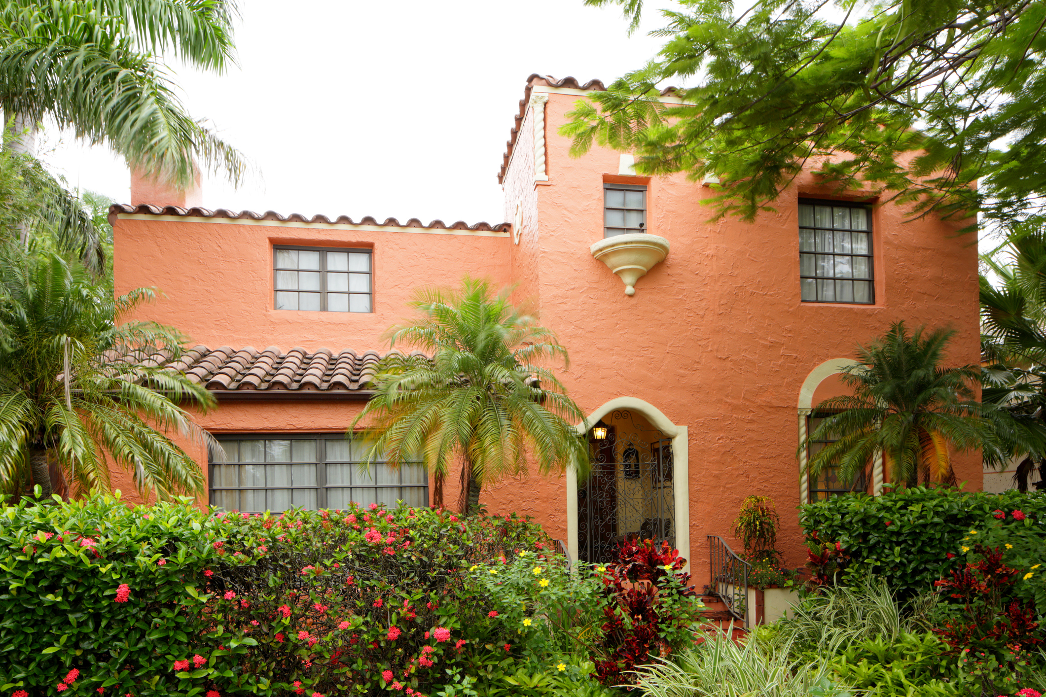The front exterior of an orange-tinted Spanish colonial style home with plants and trees in the front yard.