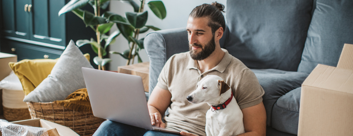 A young man looks at available houses on his laptop as his dog sits next to him.