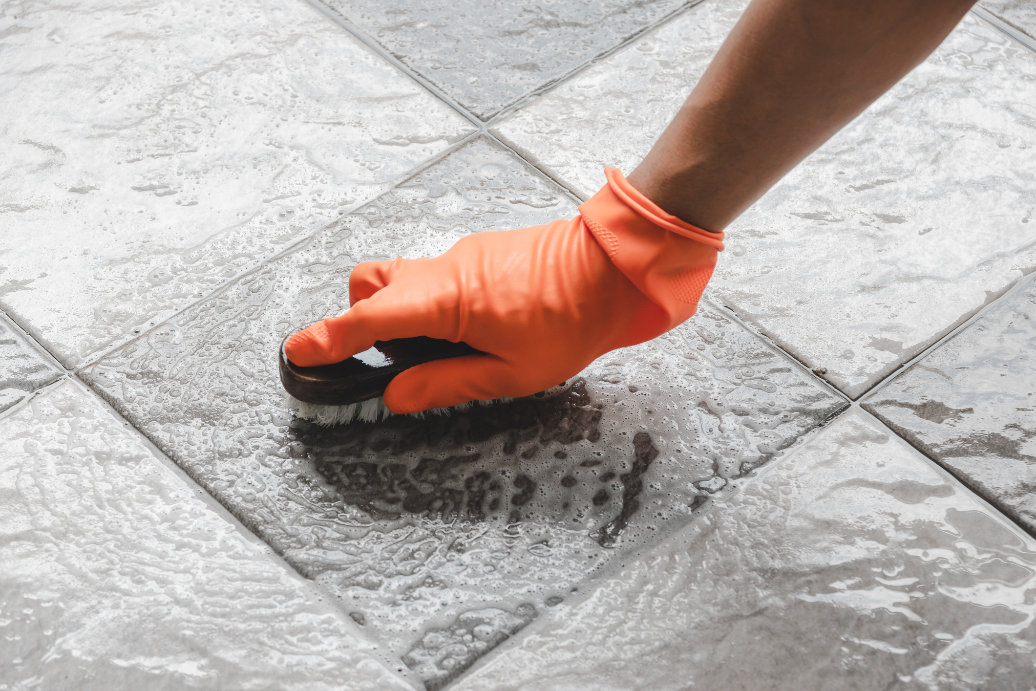 A closeup of someone scrubbing their tile floor wearing an orange rubber glove.