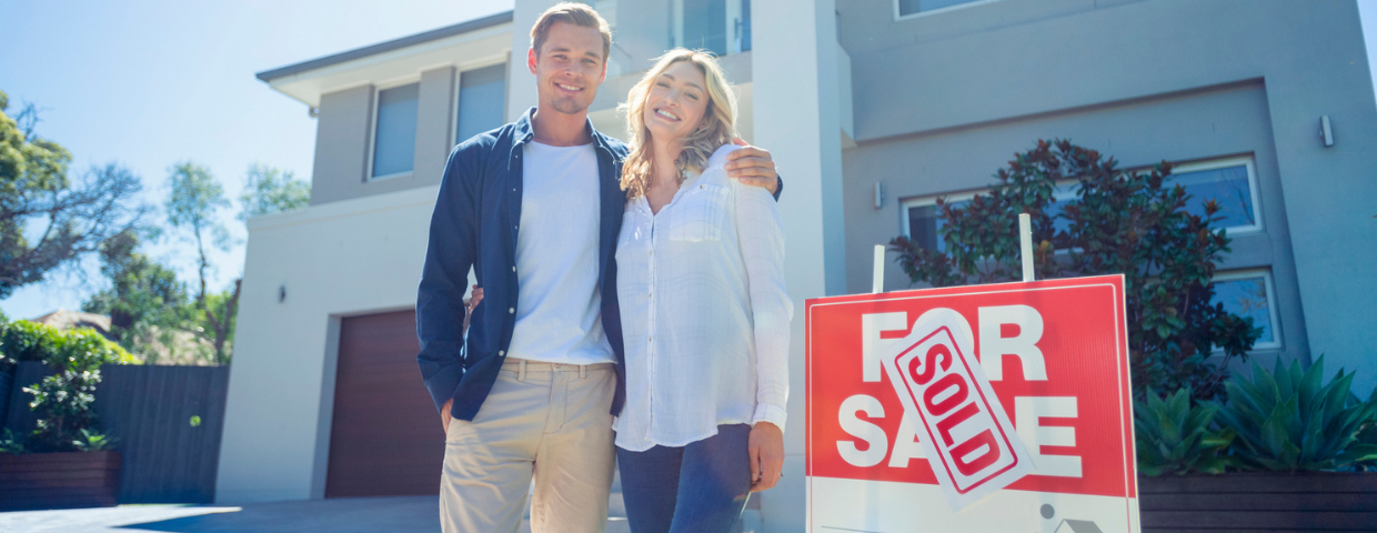 A young man and a woman stand in the driveway of their home next to a sold sign.