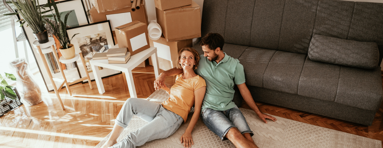 A young man and woman sit on their living room floor next to a pile of boxes.