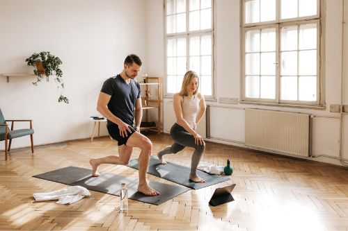 A young couple is doing lunges while watching a home workout video on a tablet. They are dressed in fashionable sportswear. They have matching dark exercise mats and exercising gear next to them. Large windows in the background bring light in their living room.