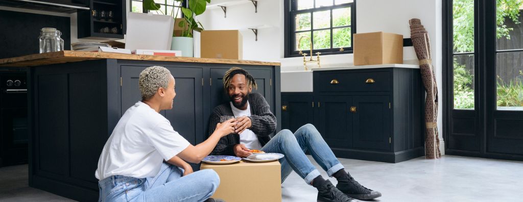 A young man and woman in the process of moving out of their home are using cardboard boxes as a table to eat their pizza as they sit on the kitchen floor