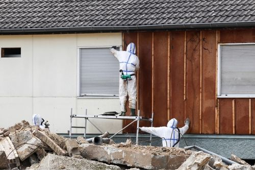 Asbestos removal professionals wearing white protective suits pry open the exterior siding of a home.