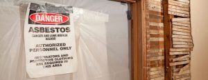 An asbestos warning sign in sealed area of a house basement with exposed sheetrock.