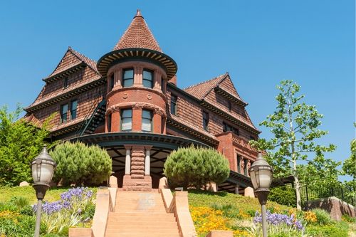 A low angle shot of a three-story brick Gothic Revival home on a corner lot with a colorful garden. The corner of the house has a round tower with a pointed roof, calling back to rounded tower castles, with a rounded wrap-around porch underneath.