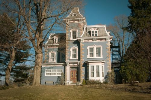 A light blue Gothic Revival home with brick trim on top of a hill on a sunny day. Corners of the home have exposed white-washed brick while the walls are light blue stucco. The windows have ornate white trim and has a double front door. The home has three sections, with the middle column the tallest, the right side is slightly protruding out and the left is tucked back behind the center tower, each has a roof with inverted gothic arches.