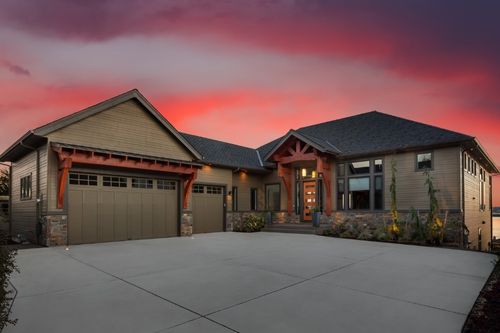 A luxury lodge-style home at sunset with timber wood framing and stone columns. The garage is attached to the home. The exterior is painted a mix of forest green and brown with deep red trim and an orange door.
