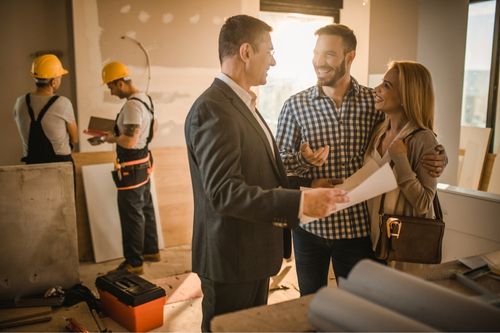 A man and woman discuss a renovation project with their real estate agent as construction contractors work in the background
