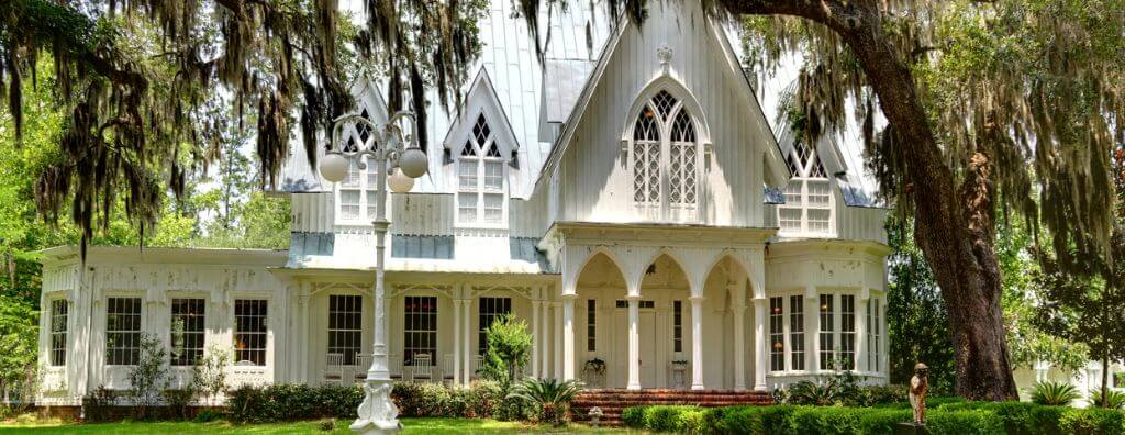 The front façade of a white Gothic Revival home in South Carolina with surrounding trees and plants. The home shows examples of Gothic Revival features, like a steeply pitched roof, arched pillars on the front porch, and church-style windows.