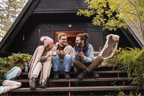 Three people enjoying a vacation on the porch of an A-Frame cabin on a cold day. Behind them, the cabin is stained black, with open French doors leading to the back porch.