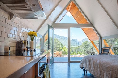 The inside of an A-Frame loft. The roof structure is wide at floor level, rising to a joined point at the top. A triangular window is constructed in the A-Frame shape, on the bottom portion of the window, a glass door is open to an open deck. In the foreground, there is a kitchen counter on the left and a comfortable bed on the right.