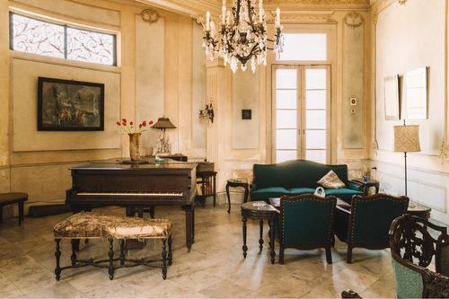 A living room with art deco decorations including a chandelier, a regal tufted green couch, gold decorations, and a dark wooden piano and coffee table.