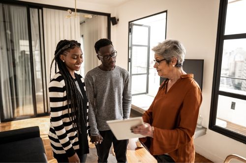 A real estate agent tours a home with two buyers. They have a brief meeting in the living room where the agent shows them paperwork for the home purchase.