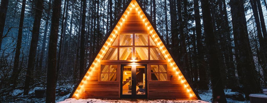 A ground-level shot of an a-frame cabin in a snowy forest. The cabin has two levels of windows, a double door, and a large front porch.