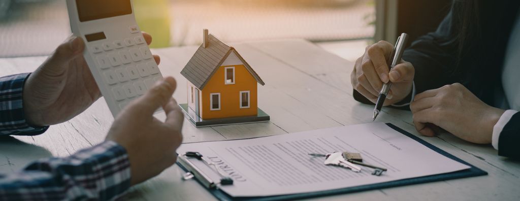 A real estate agent and a home buyer have a meeting to go over the contract of a home purchase. The real estate agent takes notes while the buyer points to a calculator as they sort through the terms of the transaction.