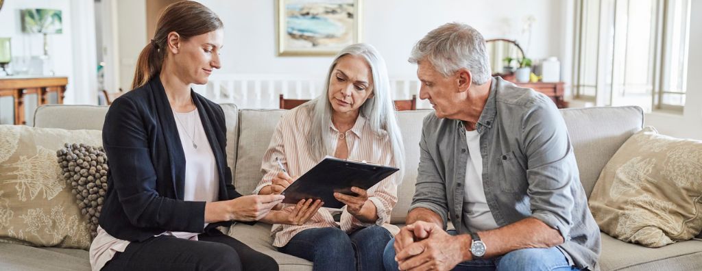 In their living room, a man and woman discuss with their real estate agent the terms of a home purchase agreement for a property under contract.