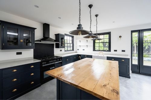 A modern kitchen with state-of-the-art appliances and matching navy-blue cabinets and drawers with gold handles. The island has a matching navy base and warm butcher-block like wood top. Against bright white walls and with metallic accents, the space is both colorful and warm.
